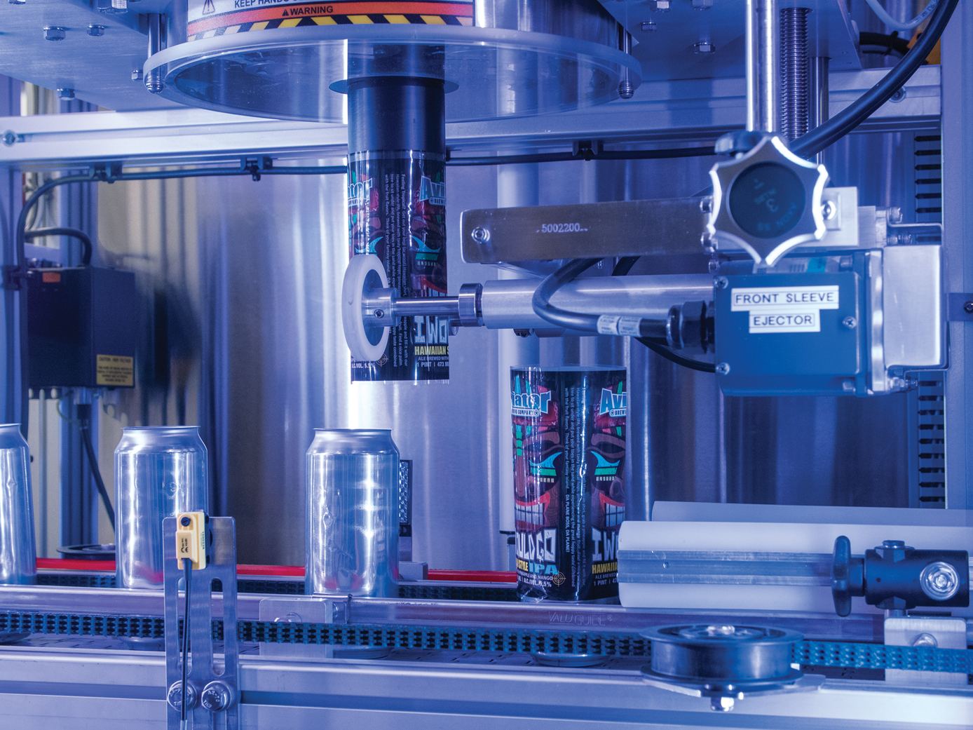 Closeup of labeler shows one can with a freshly cut label that has just been applied while the next can waits for application of its label.