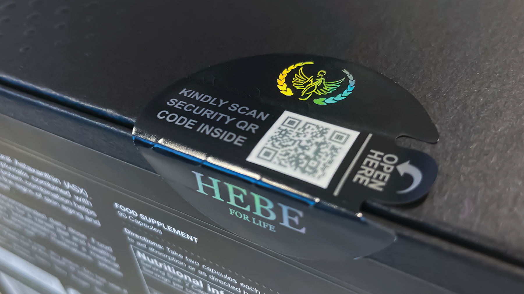 A holographically printed, tamper evident label with external QR code and die-cut tear strip spans the gap between shipper sidewalls and top.