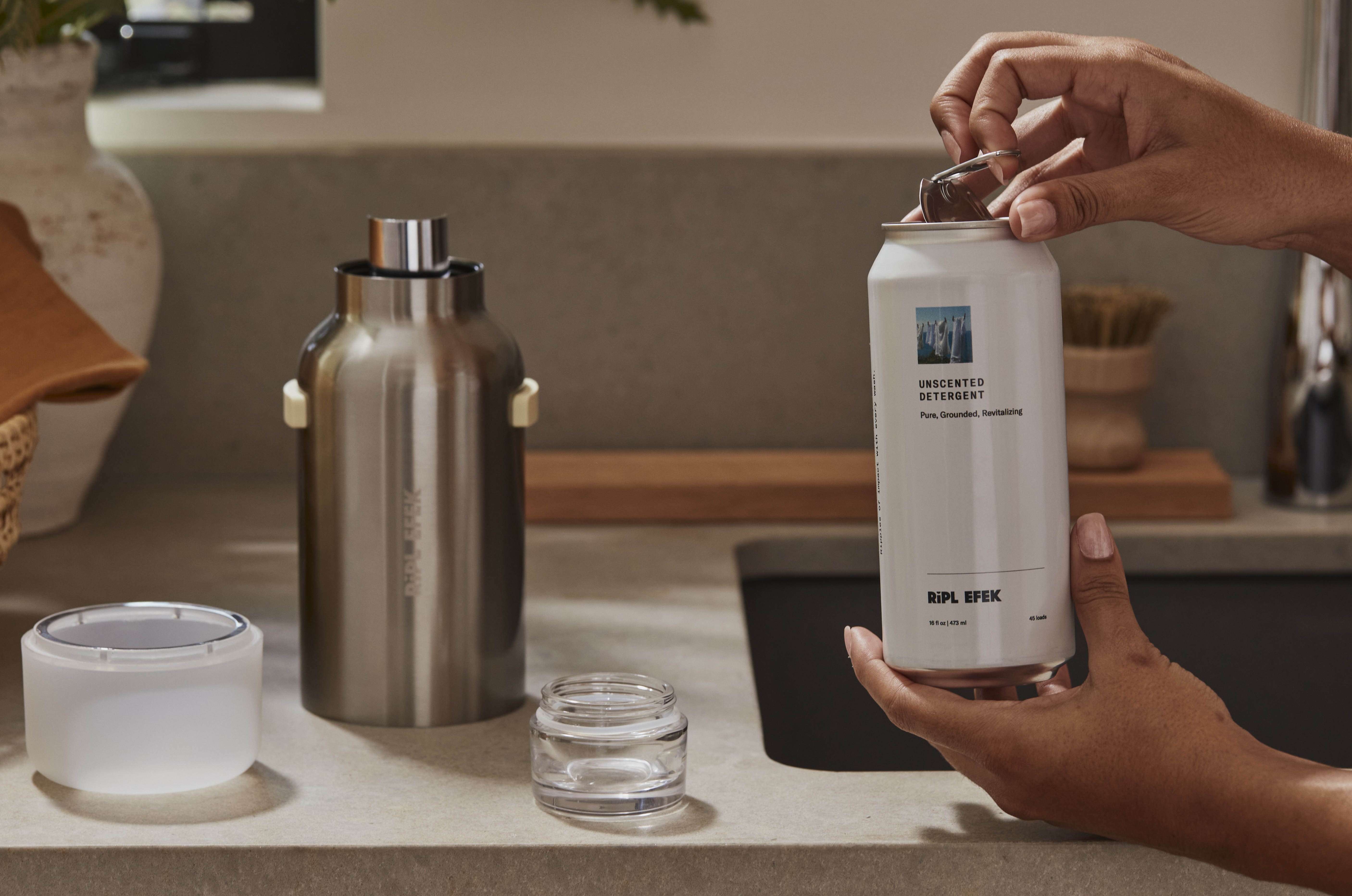 The aluminum refill can is opened and inserted into the reusable stainless steel vessel, where it locks into place for dispensing.